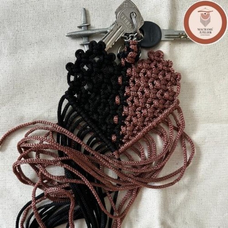 Puede incluir: Un llavero de macram&eacute; en forma de coraz&oacute;n con un dise&ntilde;o dividido, una mitad en cord&oacute;n negro y la otra en cord&oacute;n de color cobre metalizado. El llavero est&aacute; sujeto a una anilla de metal plateado que sostiene dos llaves de metal.