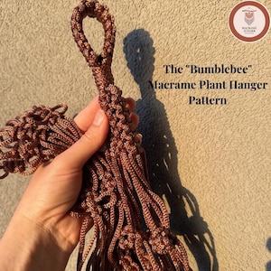 May include: A brown macrame plant hanger featuring intricate knotted cord patterns and a top loop for hanging. The text The Bumblebee Macrame Plant Hanger Pattern is visible alongside a circular logo for Macrame Atelier in the upper right corner.