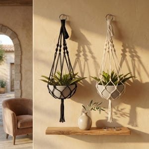 May include: Two macrame plant hangers, one black and one off-white, each holding a grey pot with a green plant. The hangers feature knotted cord designs, metal rings for suspension, and decorative tassels at the bottom, hanging against a wall.