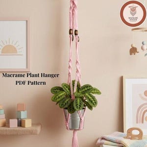 Könnte beinhalten: Ein hellrosa Makramee-Pflanzenhänger mit geknoteter Kordel, dunklen Holzperlen und einem silberfarbenen Metalltopf mit einer grün gemusterten Pflanze. Der Text Macrame Plant Hanger PDF Pattern steht in Schwarz auf der linken Seite des Bildes.