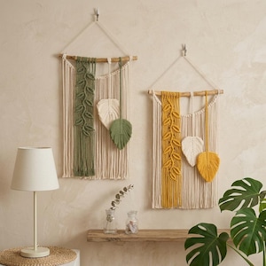 May include: Two macrame wall hangings with leaf designs. One in green and cream, the other yellow and cream. Each has a wooden dowel and hanging cord. Decorative home decor.