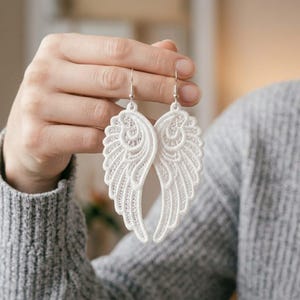 Op de afbeelding: Een paar witte kanten engelvleugel oorbellen met ingewikkeld borduurwerk en opengewerkte details. De oorbellen hangen aan zilverkleurige metalen haakjes en hebben een delicaat, lichtgewicht ontwerp met een symmetrisch verenpatroon.