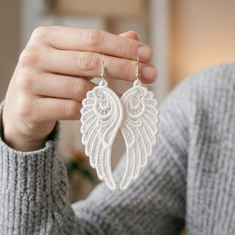 Op de afbeelding: Een paar witte kanten engelvleugel oorbellen met ingewikkeld borduurwerk en opengewerkte details. De oorbellen hangen aan zilverkleurige metalen haakjes en hebben een delicaat, lichtgewicht ontwerp met een symmetrisch verenpatroon.