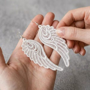 Op de afbeelding: Een paar witte kanten oorbellen in de vorm van engelvleugels. Elke oorbel is voorzien van ingewikkelde, geborduurde draadpatronen die een delicate vleugelsilhouet vormen, hangend aan een eenvoudige zilverkleurige metalen haak voor gaatjes.