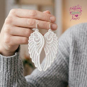 Op de afbeelding: Een paar witte kanten engelvleugel oorbellen met ingewikkelde borduurpatronen en zilverkleurige metalen haken. Het ontwerp toont gedetailleerd stikwerk dat de vorm van gevederde vleugels vormt, hangend aan eenvoudige metalen oorhaakjes.