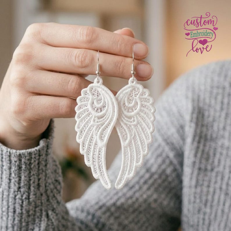 Op de afbeelding: Een paar witte kanten engelvleugel oorbellen met ingewikkelde borduurpatronen en zilverkleurige metalen haken. Het ontwerp toont gedetailleerd stikwerk dat de vorm van gevederde vleugels vormt, hangend aan eenvoudige metalen oorhaakjes.