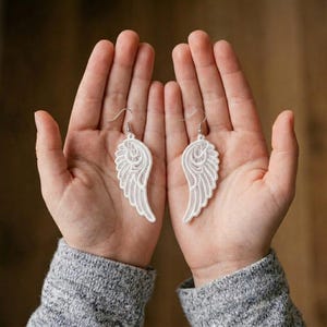 Op de afbeelding: Een paar witte kanten engelvleugel oorbellen met ingewikkelde draadpatronen en zilverkleurige metalen haakjes. De oorbellen hebben de vorm van gestileerde vogelvleugels met gedetailleerd borduurwerk en een verfijnd, lichtgewicht ontwerp.