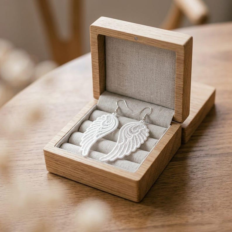 Op de afbeelding: Een paar witte, vleugelvormige kanten oorbellen met zilverkleurige haakjes, rustend in een open vierkant houten juwelendoosje met een lichtgrijze, met stof beklede binnenkant die voorzien is van meerdere gleuven voor het opbergen van sieraden.