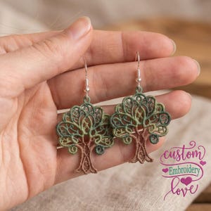 May include: A pair of drop earrings featuring intricate tree-shaped embroidery in shades of brown and green. The design showcases a detailed trunk and a sprawling canopy of stitched leaves, suspended from silver-toned metal hooks.