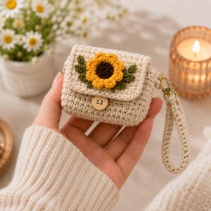 Op de afbeelding: Een beige gehaakt hoesje voor draadloze oordopjes met een klepsluiting en een enkele houten knoop. De voorkant van de klep is versierd met een gele zonnebloemapplicatie met een donkerbruin hart en groene blaadjes. Een bijpassend gehaakt polsbandje is bevestigd.