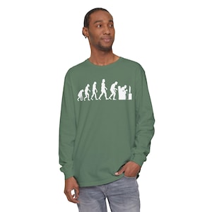 Könnte beinhalten: Ein salbeigrünes Langarmshirt mit einem weißen Grafikdruck, der die Stadien der menschlichen Evolution darstellt und mit einer Figur endet, die in einem Sessel sitzt und ein Videospiel spielt. Das Shirt hat einen Rundhalsausschnitt und lange Ärmel.