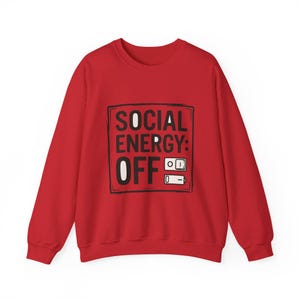 Op de afbeelding: Een rode sweatshirt met een zwarte rechthoek met de woorden "SOCIAL ENERGY: OFF" in wit en zwart. Het ontwerp bevat een aan/uit-knop en een batterijpictogram, beide in zwart en wit.