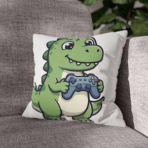 May include: A square white throw pillow featuring a cartoon illustration of a green dinosaur with dark green spots, holding a blue video game controller with colorful buttons. The dinosaur has a light cream-colored belly and a wide, smiling mouth.