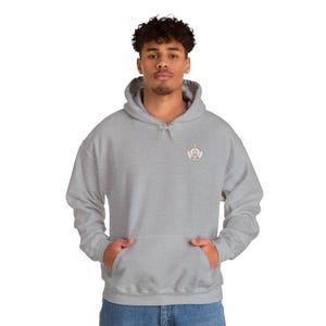 May include: A light gray hooded sweatshirt with a front pocket and a small circular emblem on the upper left chest. The emblem has text and a graphic. The model is wearing blue jeans.