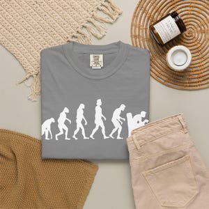 Könnte beinhalten: Ein graues T-Shirt mit Rundhalsausschnitt und einem weißen Grafikdruck, der die Evolution der Menschheit darstellt, beginnend bei einem Primaten bis hin zu einer menschlichen Figur, die auf einer Toilette sitzt. Das Shirt hat ein Comfort Colors Etikett.