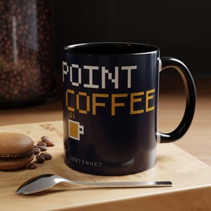 May include: A dark blue ceramic mug with a black interior and handle. The exterior features pixelated white text reading POINT and yellow text reading COFFEE, with a small yellow coffee mug icon and the white text CONTINUE? below it.