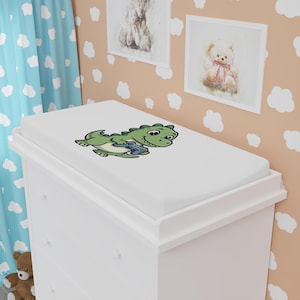 May include: A white changing pad cover featuring a cartoon green dinosaur holding a blue video game controller. The dinosaur has a light green belly, dark green spots on its back, and a friendly expression. The cover is rectangular with rounded corners.