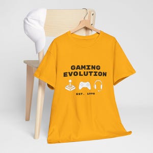 Könnte beinhalten: Ein senfgelbes T-Shirt mit kurzen Ärmeln, das den schwarzen Schriftzug GAMING EVOLUTION sowie weiße grafische Symbole eines Joysticks, eines Gamecontrollers und eines Headsets zeigt, mit dem Text EST. 1990 in einfacher schwarzer Schrift.