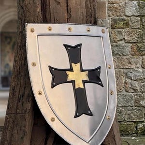 May include: A silver-colored shield with a black cross and gold accents. The shield has a rounded top and is trimmed with gold rivets. The background includes a stone wall and a wooden post.