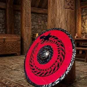 Puede incluir: Un escudo redondo rojo con un diseño de dragón negro. El escudo tiene un borde negro y plateado y un umbo central. Se apoya contra un pilar de madera en un interior rústico.