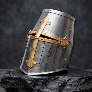 May include: A silver-toned metal great helm featuring a prominent brass-colored cross overlay on the front face. The helmet includes a horizontal eye slit, multiple small ventilation holes on the lower section, and is secured with small brass rivets.