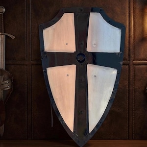 Puede incluir: Un escudo de estilo medieval con borde negro y diseño de cruz blanca. El escudo tiene un jefe central y está montado sobre una superficie marrón oscuro. Es un objeto decorativo.