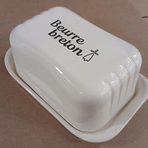 Peut inclure: Un beurrier en céramique blanc avec un couvercle, portant l'inscription "Beurre breton" en noir. Le plat est de forme rectangulaire avec un couvercle légèrement incurvé et repose sur une base assortie. Un petit motif floral accompagne le texte.