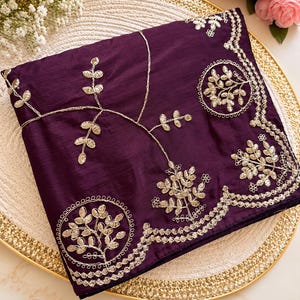 May include: A folded deep purple fabric piece featuring intricate gold metallic thread embroidery. The design includes circular motifs filled with leaf patterns and a scalloped border edge, complemented by delicate vine-like branches.