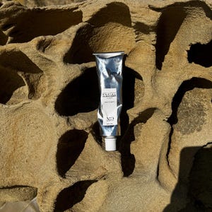 Op de afbeelding: Een zilveren tube gezichtsreiniger met witte tekst en een witte dop. De tube is gelabeld met "Facial Cleanser" en "Day/Night". Het product is 20 ml. De tube ligt op een getextureerde, bruine rotsformatie.