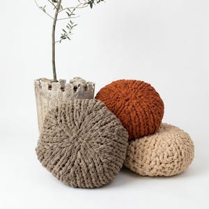 May include: Four round, knitted throw pillows in shades of brown and tan are stacked together. The pillows have a chunky knit texture. A decorative, textured, light-colored pot with a small plant is in the background.