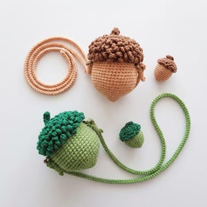 May include: Crocheted acorn-shaped purse and small acorn ornaments in shades of brown and green. The purse has a long, tan strap. The acorn tops are textured, and the purse has a detailed, knitted appearance. The background is white.