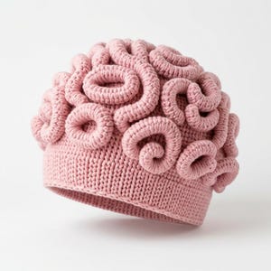 Puede incluir: Un gorro de punto rosa claro que presenta un diseño texturizado y tridimensional de bucles enroscados e interconectados en la parte superior. La base del gorro está construida con un patrón de punto acanalado estándar.