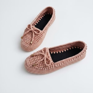 May include: A pair of handmade, light pink crochet slippers. Each slipper features a bow detail with gold-colored accents and a black interior. The slippers are designed in a moccasin style, perfect for indoor wear.