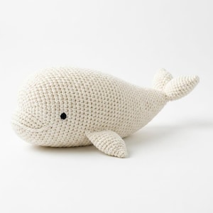 May include: A cream-coloured, crocheted beluga whale plush toy featuring a textured yarn surface, a rounded head, small side fins, a bifurcated tail, and a single small black circular eye on the side of its head, crafted with visible stitches.