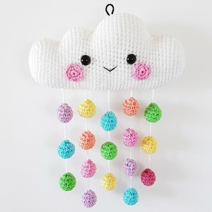 May include: A white crocheted cloud wall hanging with a smiling face, black safety eyes, and pink cheeks. Five white strings hang from the bottom, each featuring small, colorful crocheted spheres in shades of yellow, pink, blue, and green.