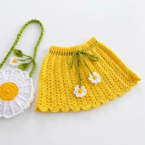 Pode incluir: Uma saia amarela de crochê com bainha ondulada e laços verdes adornados com detalhes de margaridas brancas. Uma bolsa combinando em forma de margarida com uma alça verde e folhas também é visível, criando um conjunto coordenado.
