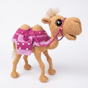 Puede incluir: Un camello de ganchillo color canela con una manta de silla de montar rosa decorada con cuentas blancas, una brida rosa a juego, grandes ojos de plástico negro y un mechón de hilo claro en la cabeza. El camello tiene dos jorobas y cuatro patas.