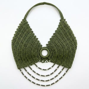 May include: An olive green crochet halter top featuring a central circular ring detail. The garment is embellished with four cascading strands of alternating small white pearls and dark metallic beads hanging from the bottom edge of the ring.