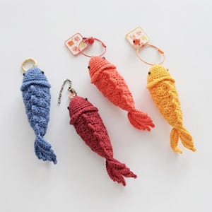 Puede incluir: Un conjunto de cinco llaveros de peces de ganchillo en varios colores: azul, rojo, naranja y amarillo. Cada pez tiene un patrón de escamas detallado, un pequeño ojo negro y una aleta caudal. Algunos llaveros tienen un anillo o cadena de metal.