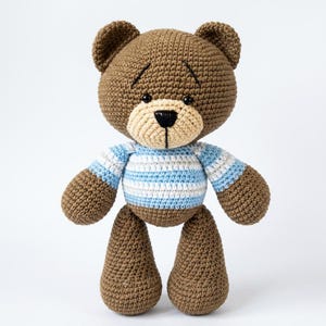 May include: A handmade brown crochet teddy bear with a light tan muzzle and a black nose. The bear wears a blue and white striped shirt. The bear has small black eyes and is standing upright.