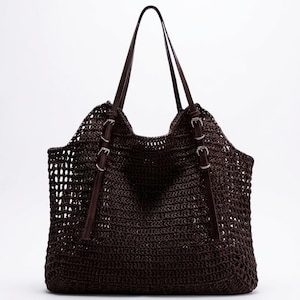May include: A dark brown tote bag featuring an open-weave crochet construction. The bag includes two long shoulder straps and two decorative vertical straps on the front, each secured with a silver-toned metal buckle for an adjustable appearance.