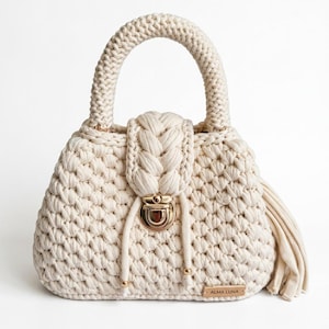 May include: A cream-colored crocheted handbag featuring a thick braided top handle and a matching braided flap closure. The bag includes a gold-toned metal push-lock clasp, two hanging cord accents with gold beads, and a small rectangular tag labeled ALMA LUNA.