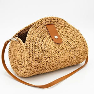 May include: A semi-circular woven straw handbag in a natural tan colour. The bag features a brown faux leather strap and a matching brown faux leather tab closure secured with a silver-toned metal snap button at the centre of the curved top.