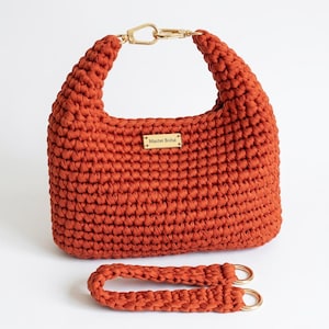 Puede incluir: Un bolso de ganchillo color óxido con un patrón de punto grueso y texturizado. El bolso incluye un cierre de metal dorado en la parte superior y una placa rectangular dorada con el texto Master Bolsa. Una correa de ganchillo a juego con anillos dorados se encuentra debajo.
