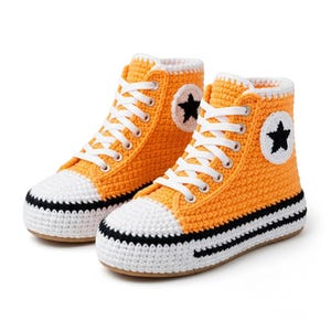 May include: A pair of high-top sneakers crafted from orange and white yarn. The shoes feature white laces, a white toe cap, a black horizontal stripe along the sole, and a circular white patch on the side with a black star in the center.
