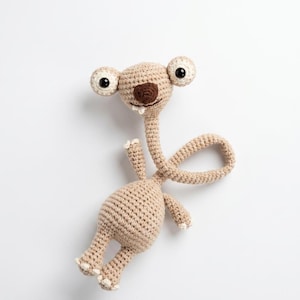 Può includere: Un animale di peluche lavorato all'uncinetto color beige con un collo lungo, un corpo arrotondato, due piccole braccia e due gambe. La testa ha due grandi occhi bianchi con pupille nere, un naso marrone e due piccoli denti bianchi sporgenti.