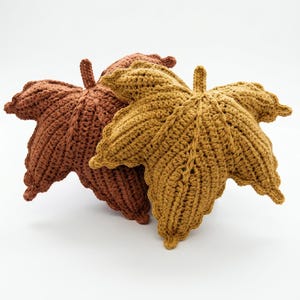 Op de afbeelding: Twee decoratieve kussens in de vorm van esdoornbladeren, één in diep roestbruin en de andere in goudkleurig mosterdgeel. Beide items hebben een getextureerd, handgehaakt oppervlak met zichtbare verhoogde nerven vanuit de centrale steel.