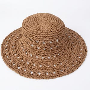 May include: A brown woven sun hat with a wide brim and a rounded crown. The hat features decorative cut-out patterns around the brim. The hat is made of a natural fiber material, perfect for sun protection and a stylish accessory.