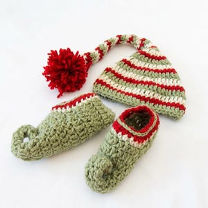Peut inclure: Un ensemble de Noël fait à la main comprenant un chapeau d'elfe crocheté avec un pompon rouge et un motif rayé vert et rouge, ainsi que deux chaussons d'elfe verts avec des bordures rouges et blanches.