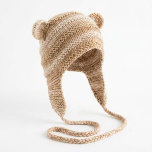May include: A knitted hat in shades of tan and cream, featuring small, rounded ear flaps and two small, rounded ears on top. The hat has ear flaps that extend down and are connected by a braided tie. The hat is designed to keep the head and ears warm.